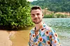 Ung mand i blomstret skjorte står og smiler ved en tropisk strandkant