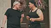 Torben og Michael fra 'Gift ved første blik' på bryllupsrejse i sort tøj taler sammen foran en dør med blomsterkranse og lyserøde bånd.