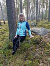 Kvinde i turkis jakke sidder på mosbegroet sten i åben fyrreskov