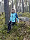 Kvinde i turkis jakke sidder på mosbegroet sten i åben fyrreskov