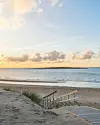 Trærampe ned gennem klitter til rolig sandstrand og hav ved solnedgang.