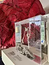 Glasmonteret skulptur foran stor rød tekstilkunst på museumssokkel.