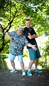 To mænd i sommeroutfit står og poserer smilende på en sti i en grøn park.