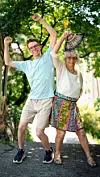 Ældre kvinde og yngre mand danser med løftede arme på en solrig sti i en grøn park.