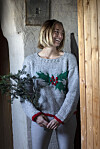 Kvinde i grå juletrøje med kristtorn står i døråbning og holder grønne grene.