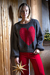 Kvinde i grå sweater med rødt hjerte og røde bukser står ved trævæg med julestjerne.