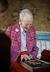 Dronning Margrethe i farverig jakke ser ned i en stor illustreret bog i stuen