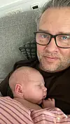 Voksen med briller holder sovende spædbarn på brystet i sofa