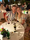 Markus og Cecilie smiler ved bord på stemningsfuld restaurant med vinflasker og blomster