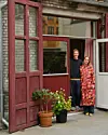 To personer står smilende i en åben dør med røde træporte og potteplanter foran.
