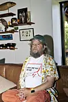 Anders Lund Madsen med kasket og trykt T-shirt sidder på brun lædersofa foran væg med hylder og dekorationer.