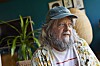 Anders Lund Madsen med skæg og kasket sidder i en farverig stue med plante bagved.