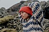 Person i rød hue og mønstret sweater står foran mosdækkede klipper