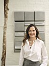 Louise Herping Ellegaard i hvid skjorte foran gråt geometrisk kunstværk på lys mur