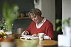 Ældre kvinde læser en roman ved spisebordet med en kaffekop foran sig.