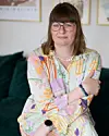 Kvinde med briller og farverig kjole sidder på mørkegrøn sofa foran væg med billeder.