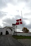Dannebrog på halv stang foran hvidkalket kirkeport en overskyet dag