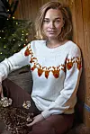 Kvinde i hvid julesweater med rensdyr sidder ved et oplyst juletræ indendørs