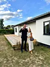 To personer står foran et hvidt hus med træterrasse i en have.