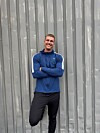 Mand i sportstøj står smilende foran en grå bølgepladevæg.