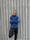Mand i sportstøj står smilende foran en grå bølgepladevæg.