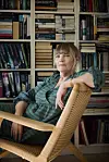Anna Grue sidder i fletlænestol foran en bogreol fyldt med bøger